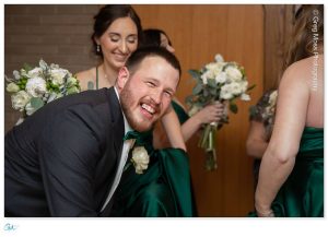 A joyful man in a suit smiles broadly at Stephanie and Corey's wedding, with women in green dresses holding bouquets in the background.