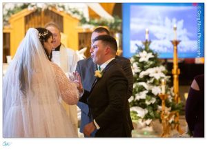 Stephanie and Corey holding hands during their wedding ceremony at Mill 1 in Holyoke, MA, with a priest officiating in the background.