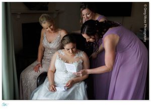 A bride in a white gown reads a letter while surrounded by three women in elegant dresses, expressing emotional support at a Holyoke Wedding Photography session.