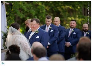 Bride and groom standing at the altar during a Highfield Hall wedding ceremony, with the groom looking at the bride affectionately, surrounded by groomsmen in blue suits.