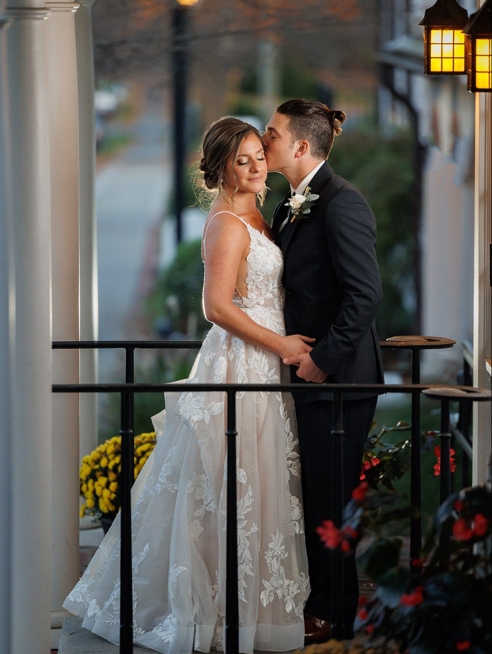 A bride and groom share a kiss on a balcony adorned with flowers, with streetlights and trees in the background at dusk, captured by Western Ma Wedding Photography.