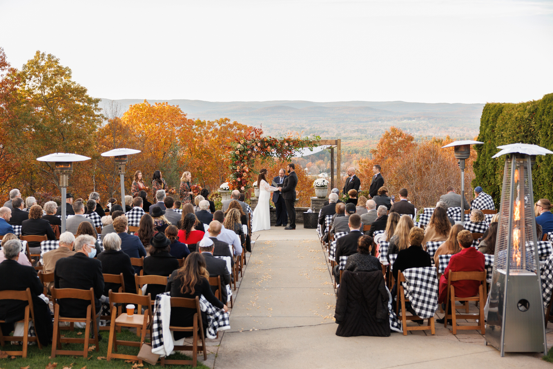 An outdoor wedding ceremony with guests seated facing a couple at the altar, set against a backdrop of autumn hills, perfectly planned for exquisite wedding photography.