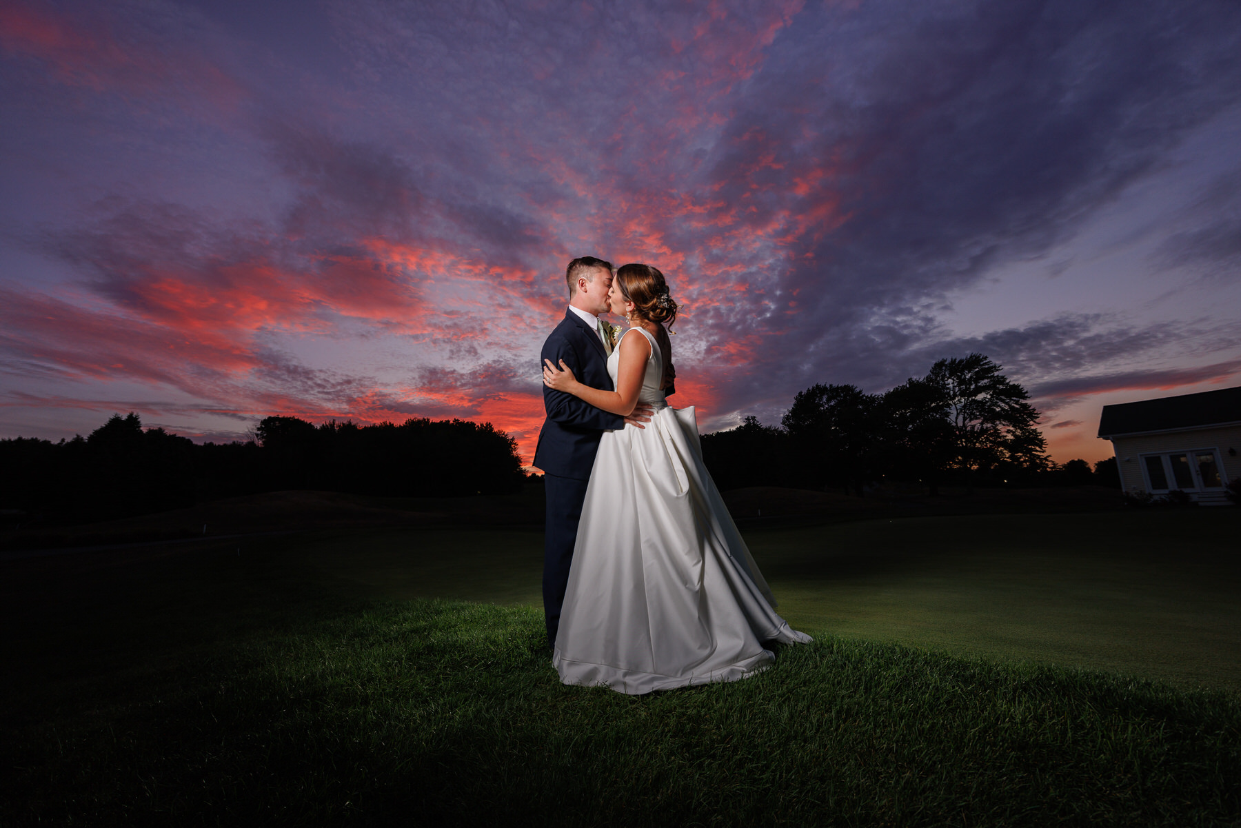 A couple kisses on a golf course at sunset, with a vibrant pink and blue sky above and a white building in the background, perfectly capturing their wedding timeline.