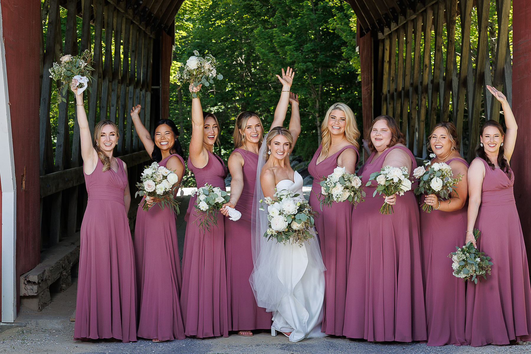 A bride and her bridesmaids, all in burgundy dresses except the bride in white, joyfully raising bouquets inside a wooden covered bridge, capturing optimal photography moments.