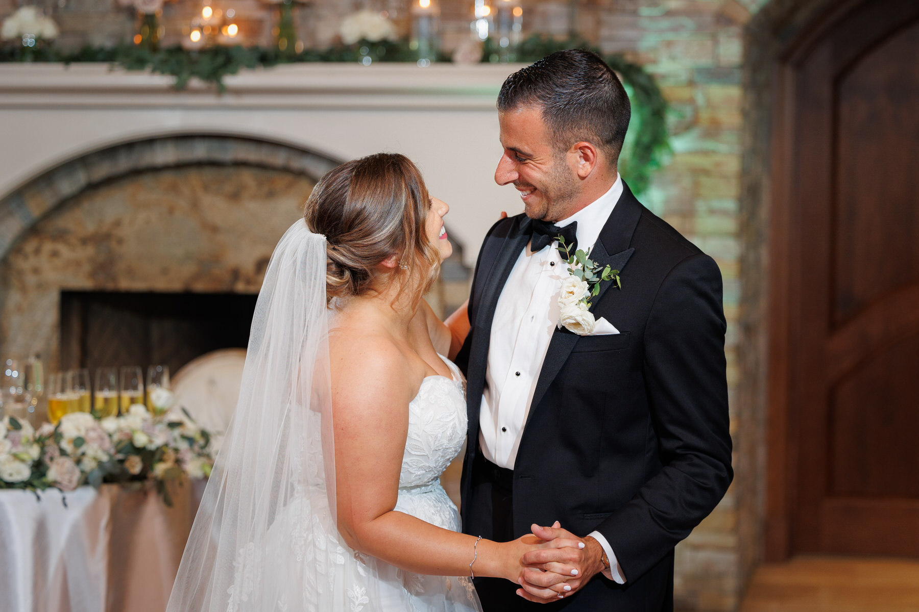 A bride and groom smiling at each other inside a room, with a decorated fireplace and floral arrangements in the background, perfectly arranged according to their wedding timeline.