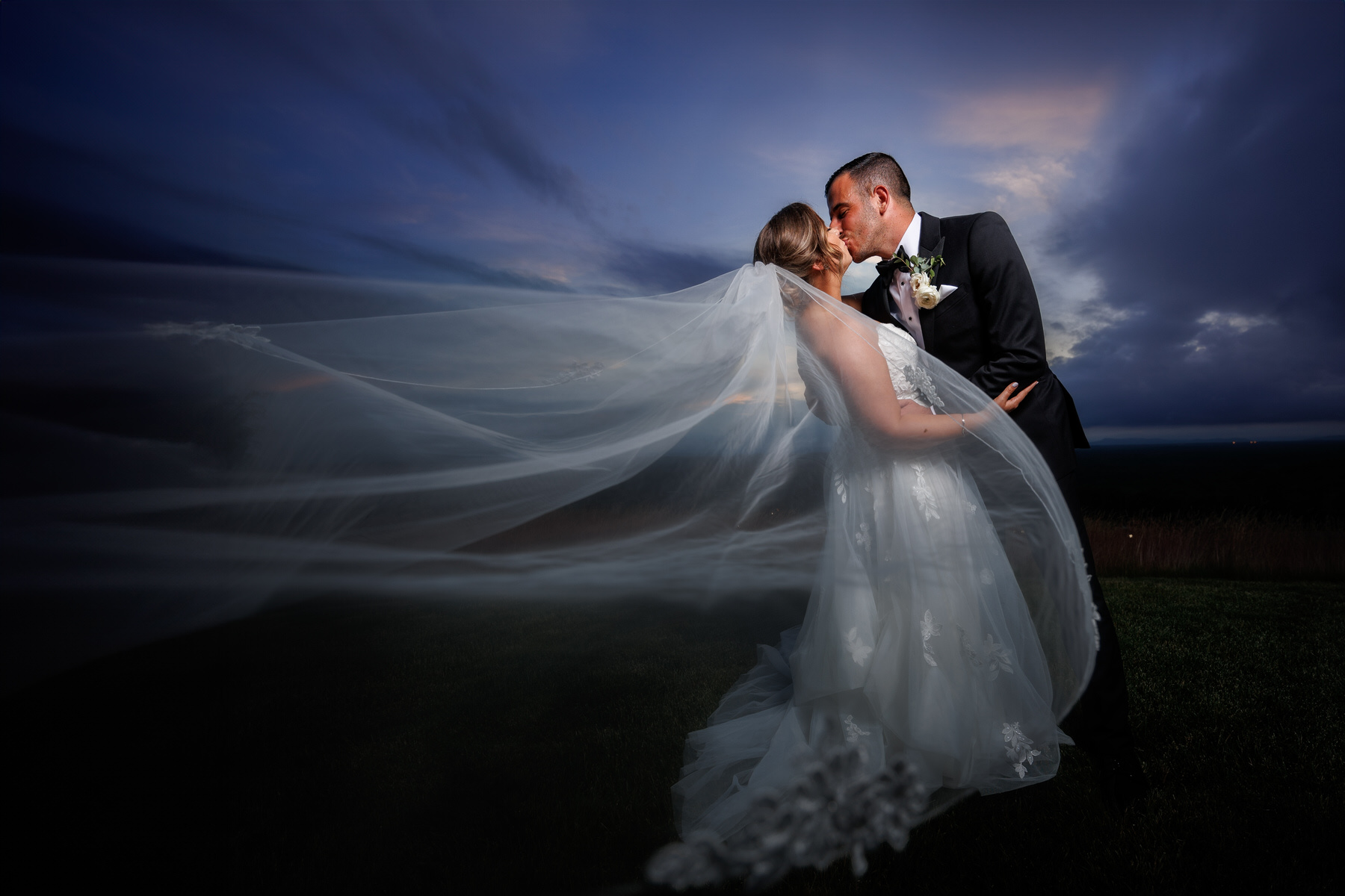 Bride and groom kissing outdoors at dusk, with the bride's long veil elegantly blowing in the wind under a dramatic cloudy sky, captured perfectly in their wedding photography.