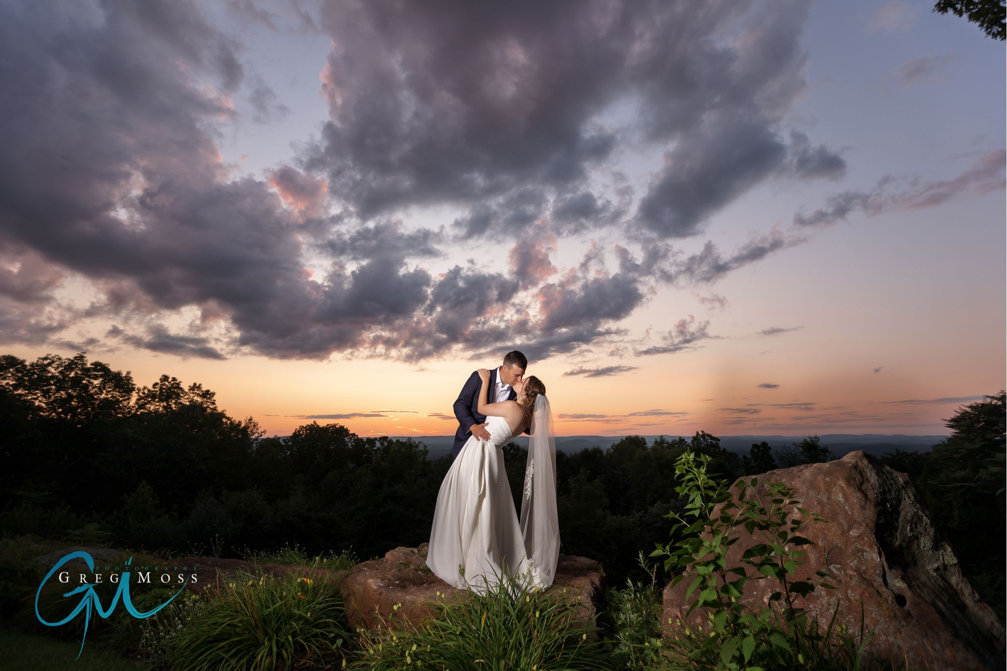 A bride and groom embracing on a rocky overlook with a vibrant sunset and dramatic clouds in the background, perfectly timed for optimal photography.