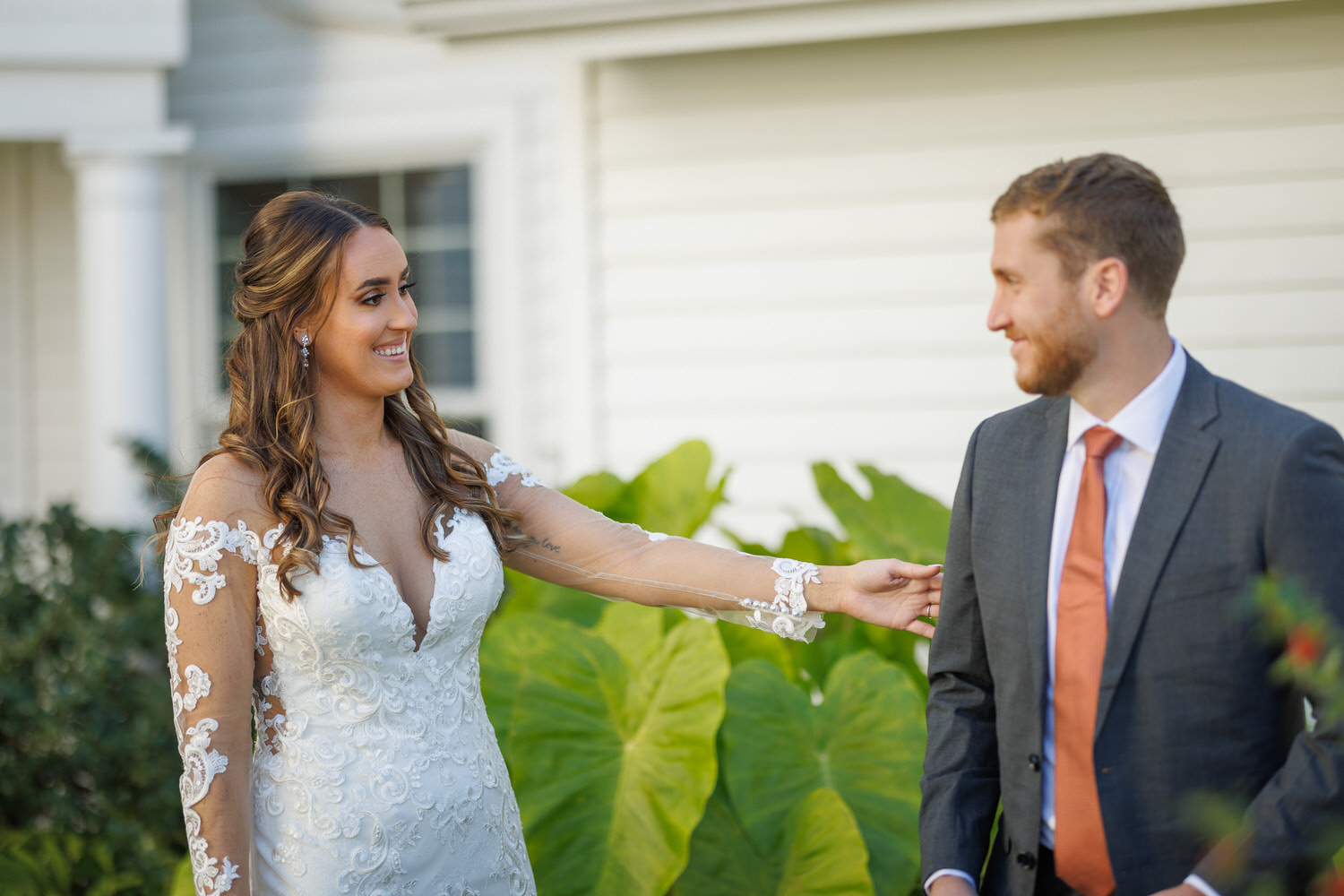 Bride in a lace dress smiling at the groom in a suit, pinning a flower on his lapel outdoors during their wedding photography session, with a white house in the background.