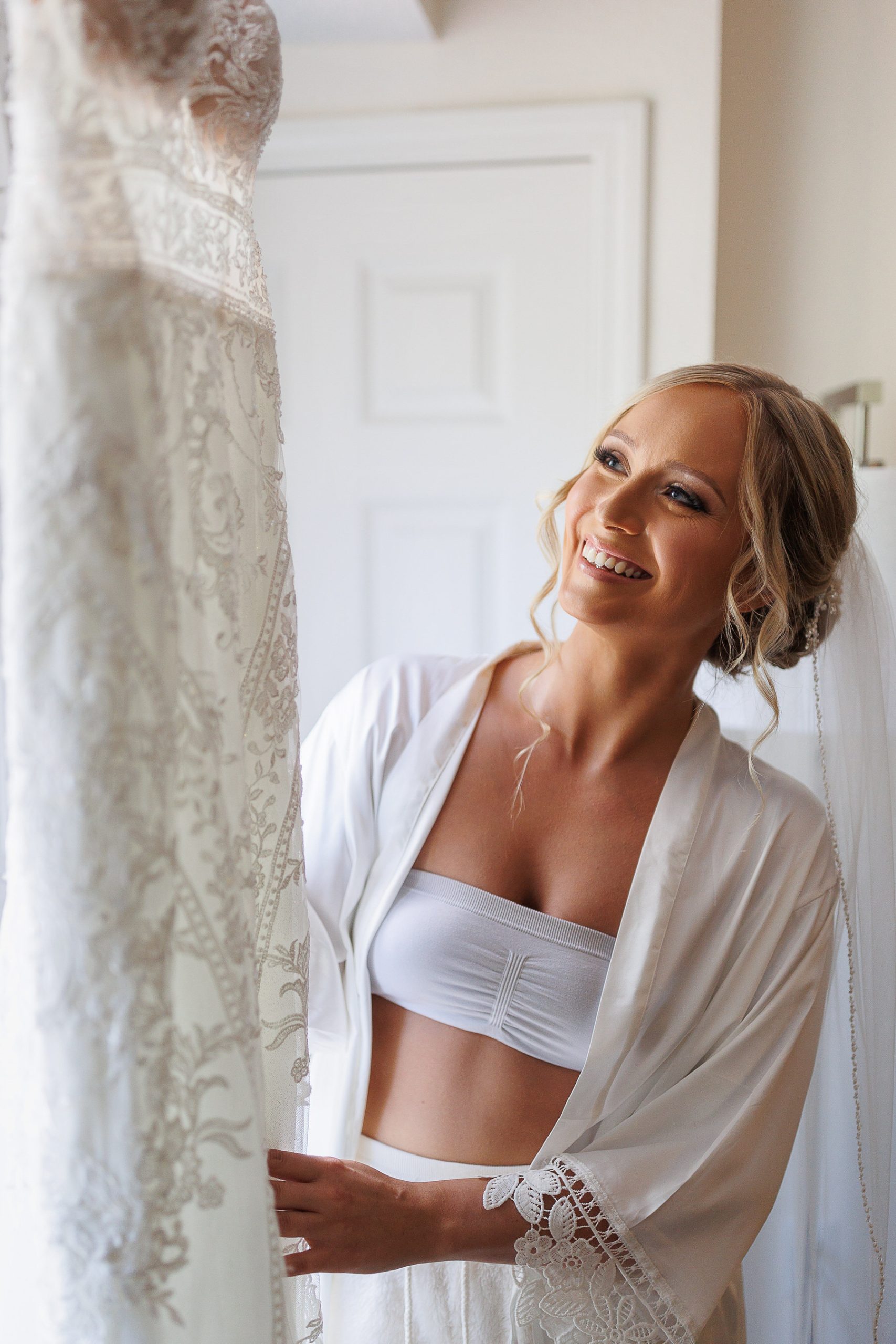 A bride in a white robe smiles as she looks at her wedding dress hanging by the door, envisioning the moments that will soon be captured in her wedding photography.