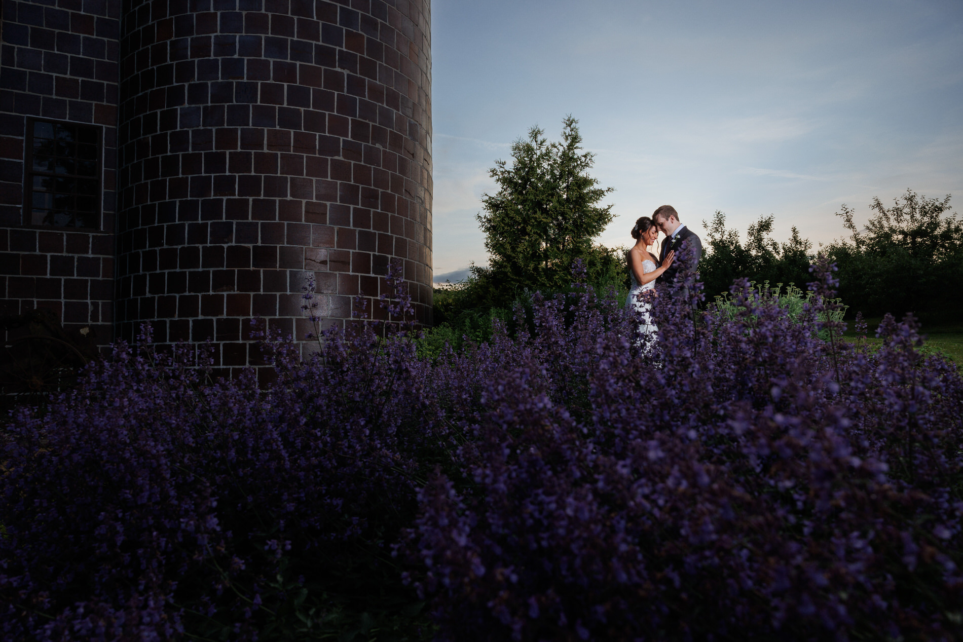 A romantic image of a couple embracing among purple flowers at twilight, with a distinctive, cylindrical brick building in the background, evoking the charm of Western Massachusetts wedding venues.