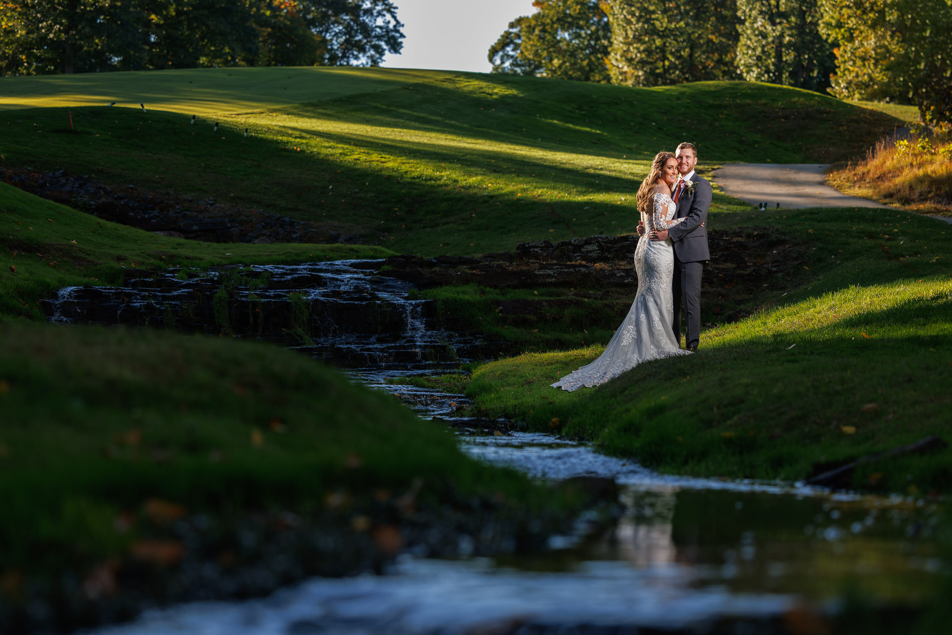A newlywed couple embraces beside a small stream with a cascading waterfall in a lush, sunlit park in Western Massachusetts.